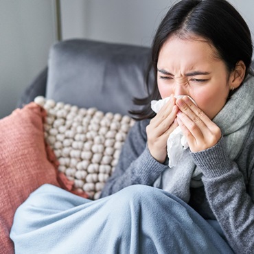 Frau sitzt auf Sofa und schnäuzt sich die Nase mit einem Taschentuch.