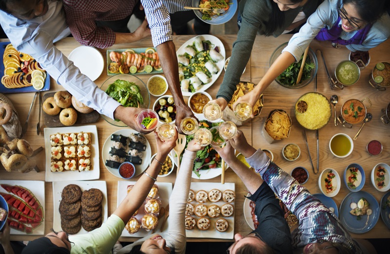 Menschengruppe sitzt gemeinsam an Tisch mit großer Auswahl an Essen und Getränken.